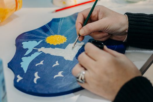 Close-up of hands painting a blue shield with birds, clouds, and sun.